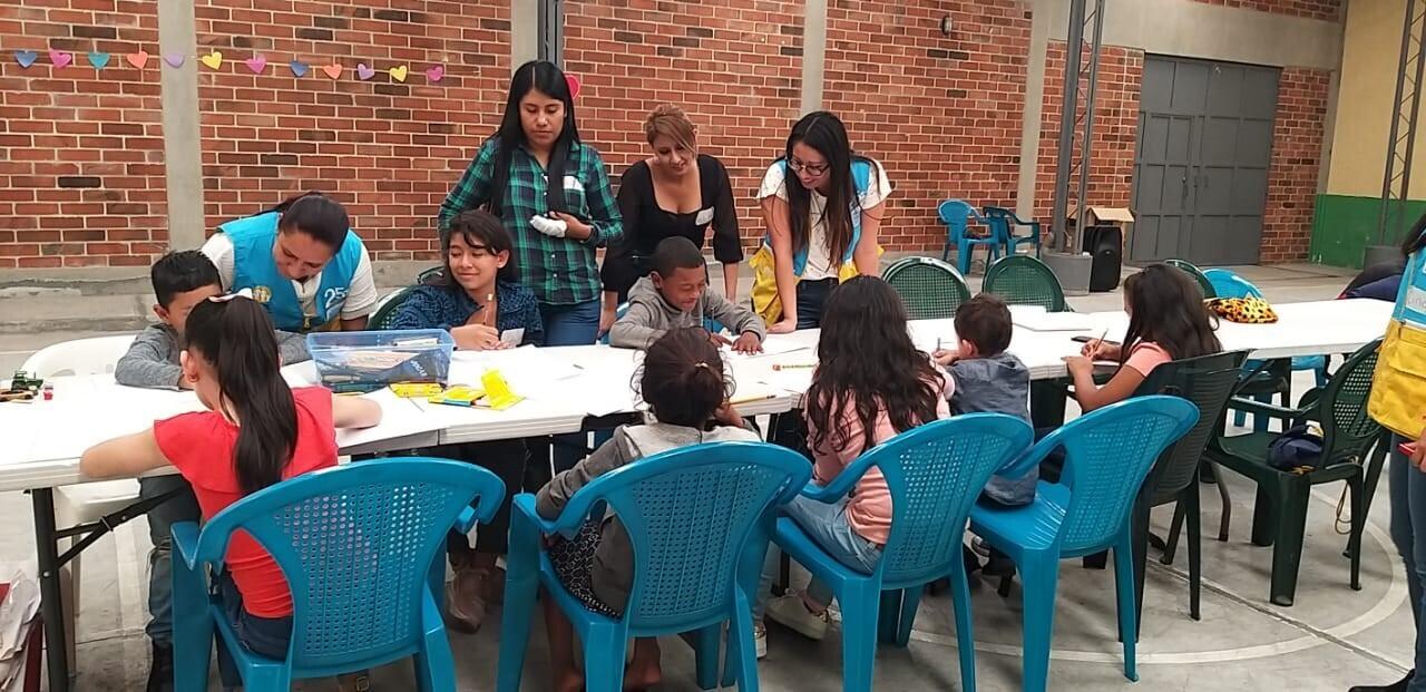 A group of women work with a group of children who are sitting and drawing at a table.