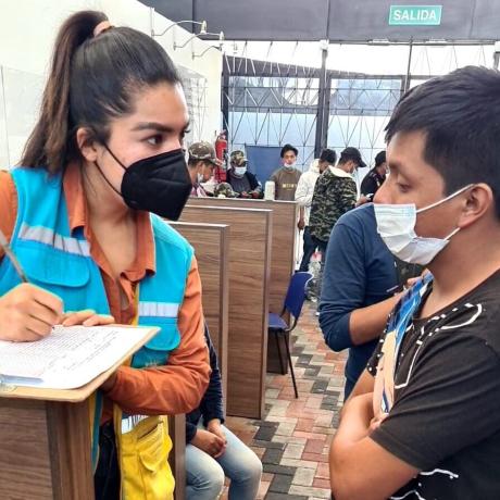 A young Latinx woman in a blue vest and face masks interviews a young man in a face mask. She is writing on a clipboard.