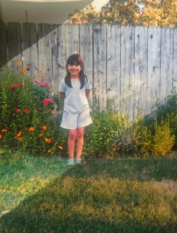 A young Latinx girl stands in a yard in front of flowering bushes and a wooden fence.