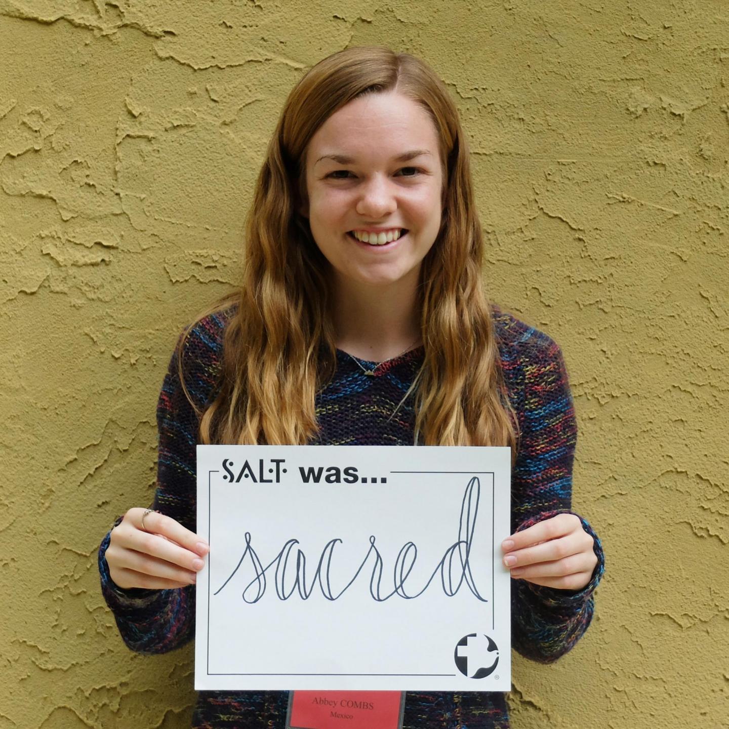 A young adult stands in front of a yellow wall and holds a sign that says, "SALT was sacred"