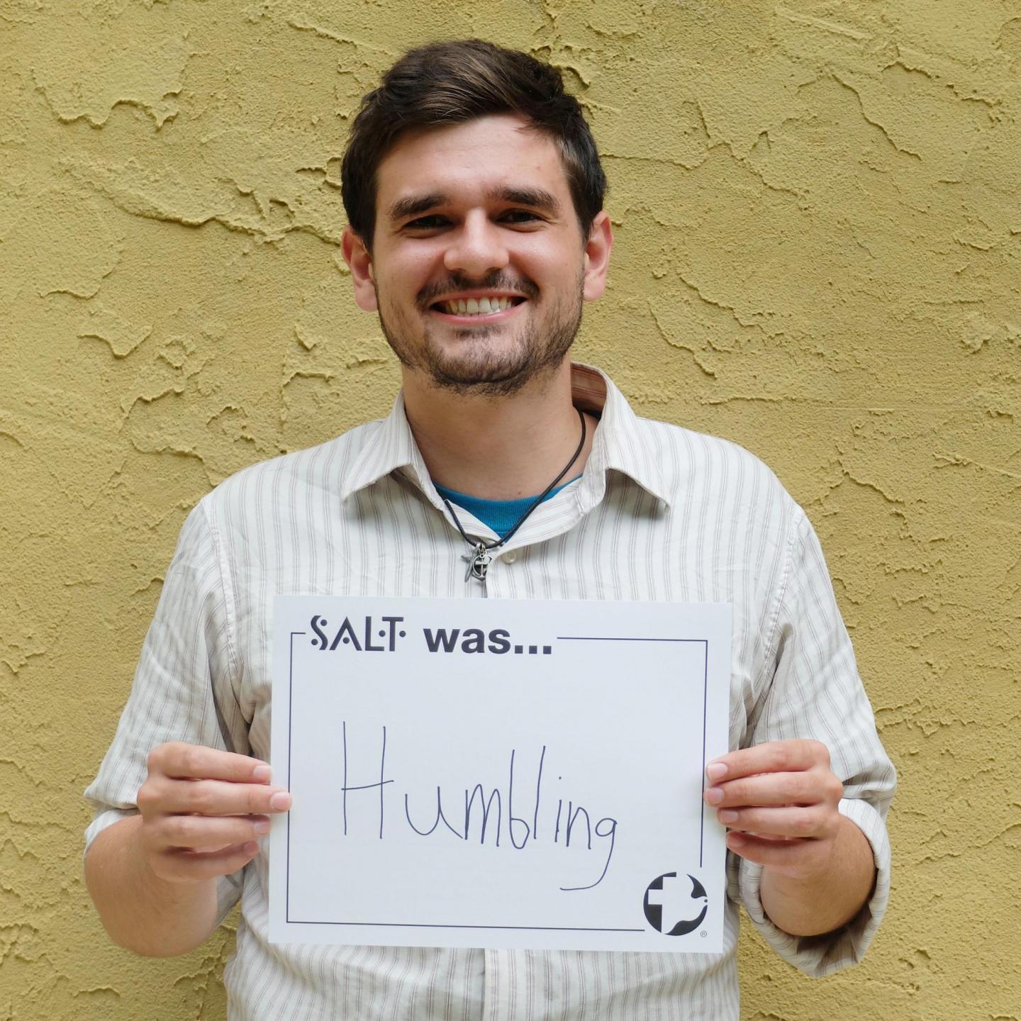 A young adult stands in front of a yellow wall and holds a sign that says, "SALT was humbling"