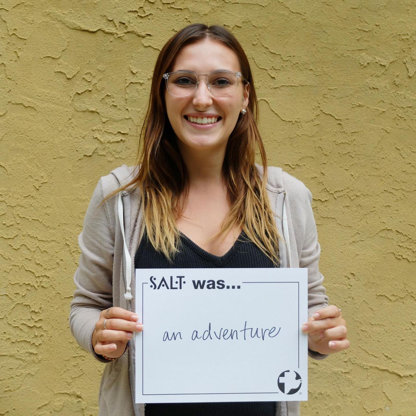 A young adult stands in front of a yellow wall and holds a sign that says, "SALT was an adventure"