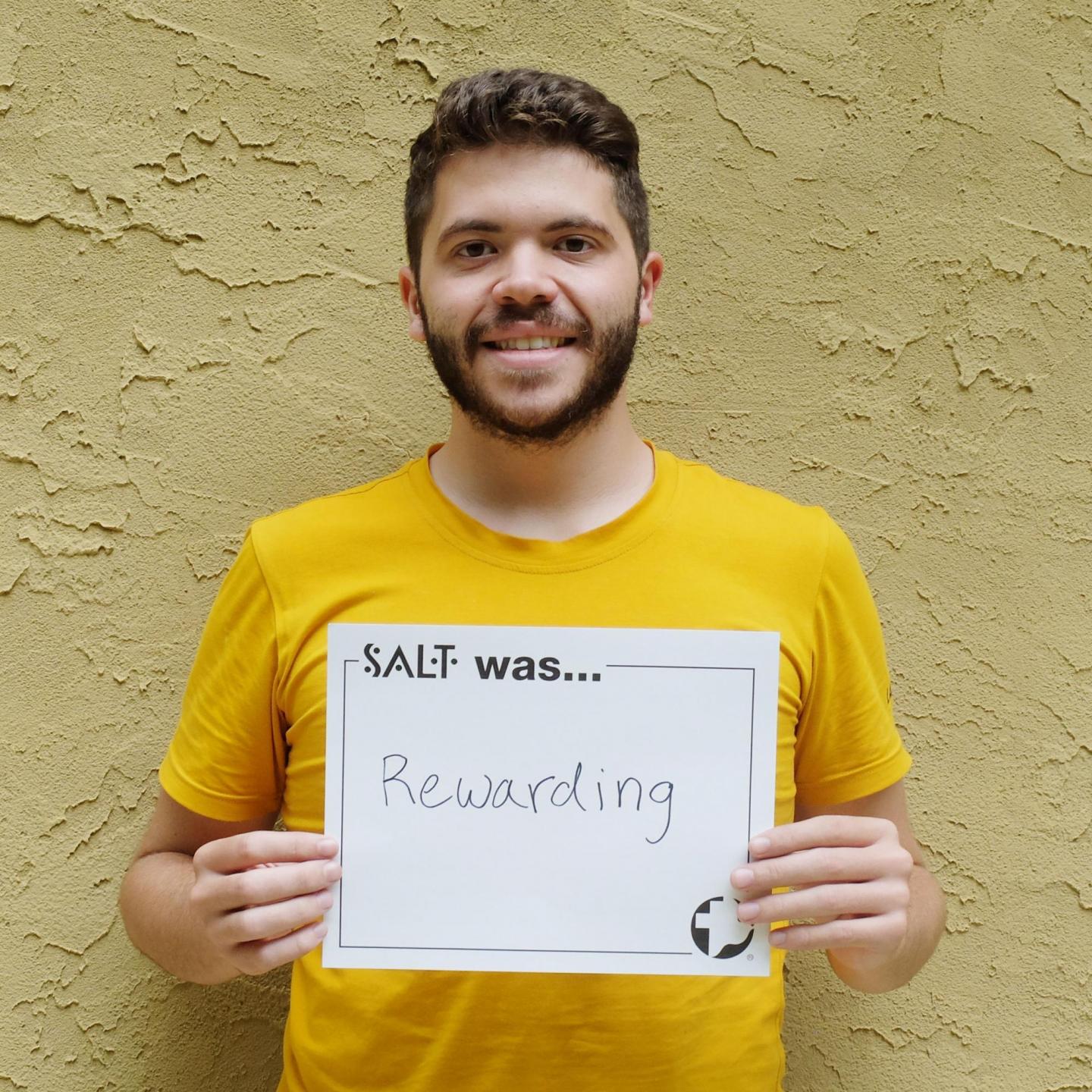 A young adult stands in front of a yellow wall and holds a sign that says, "SALT was rewarding"