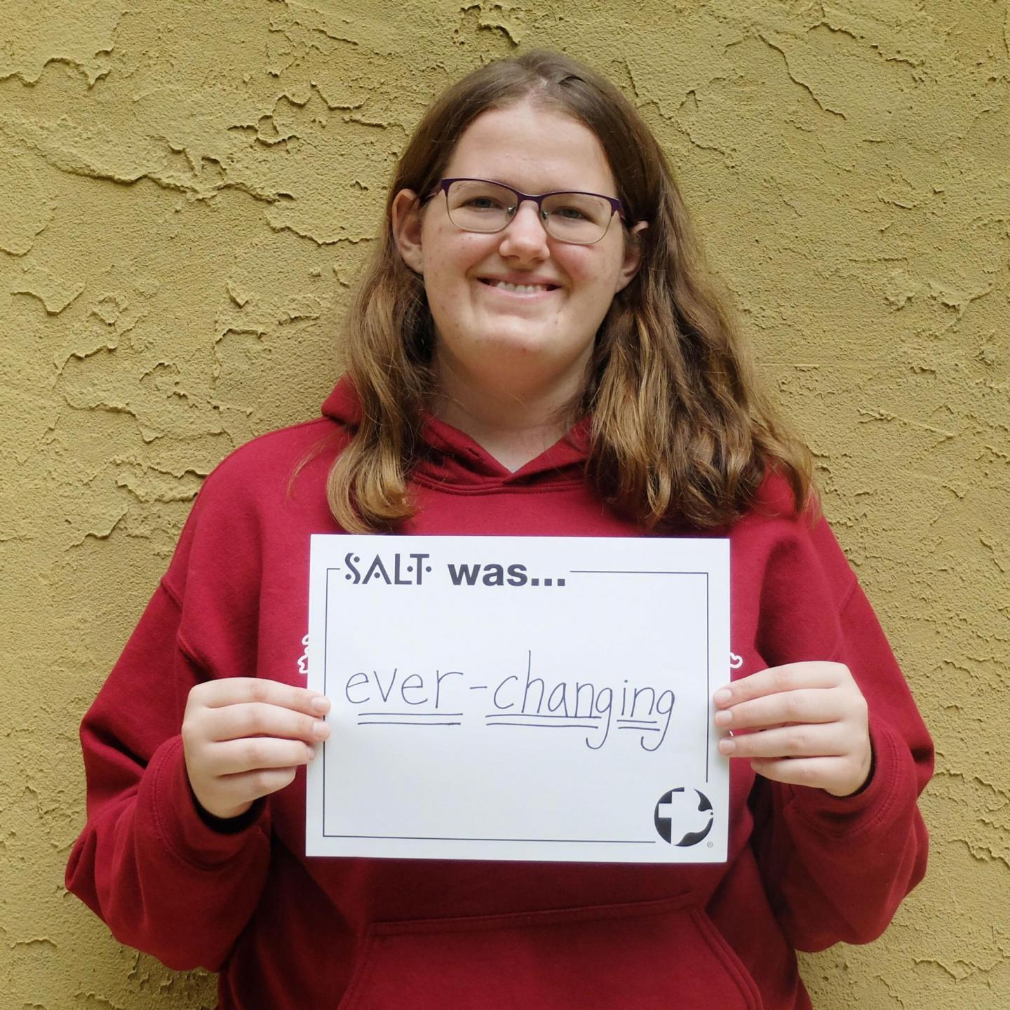 A young adult stands in front of a yellow wall and holds a sign that says, "SALT was ever-changing"