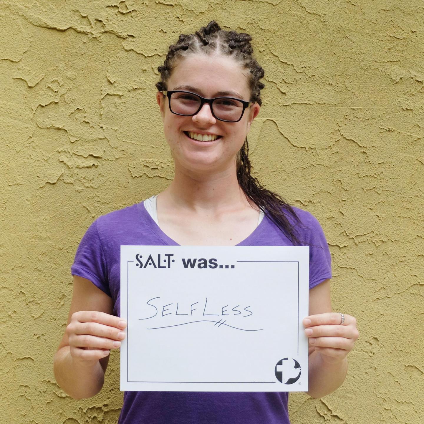 A young adult stands in front of a yellow wall and holds a sign that says, "SALT was selfless"