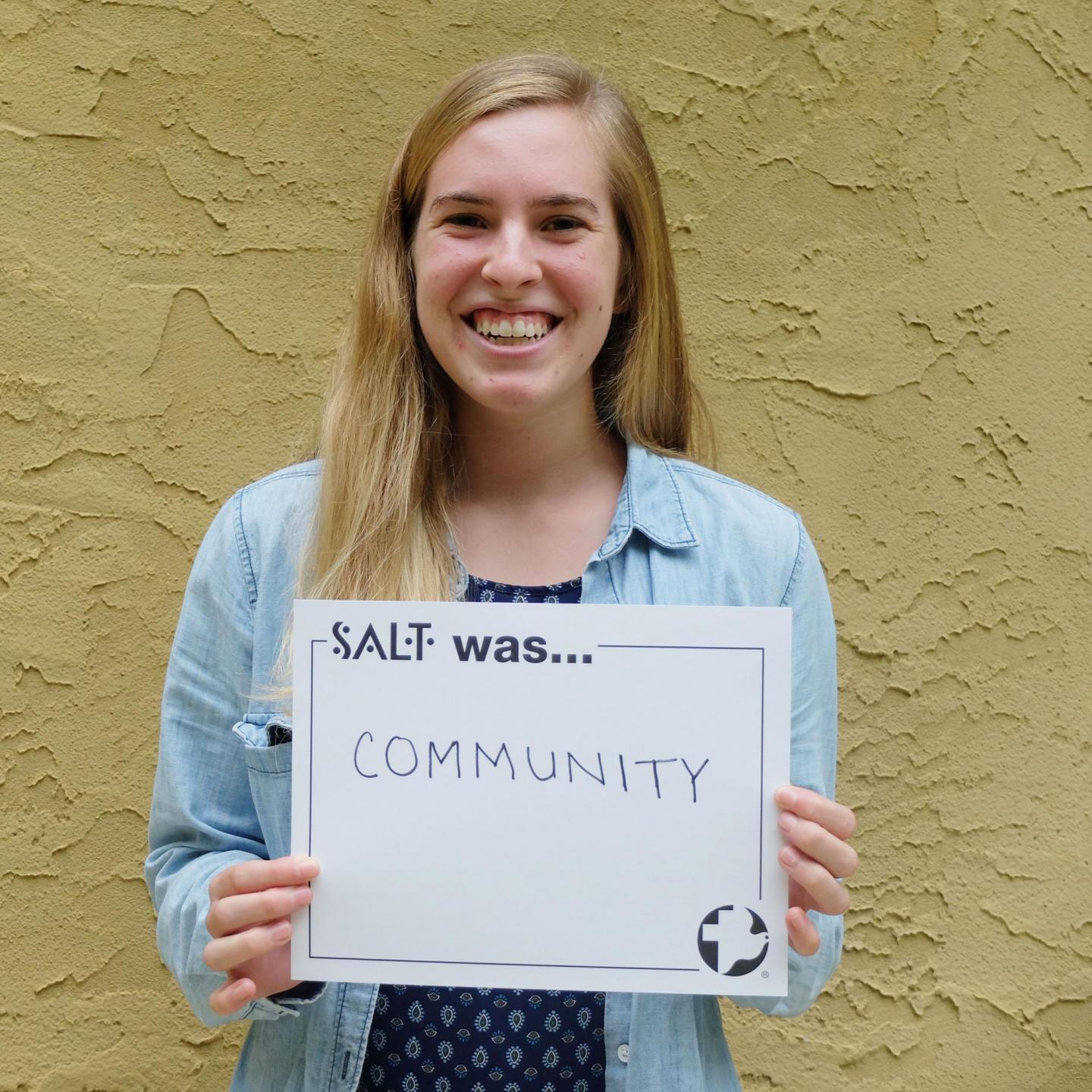 A young adult stands in front of a yellow wall and holds a sign that says, "SALT was community"