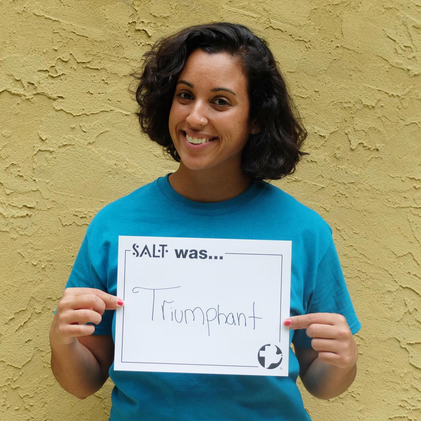 A young adult stands in front of a yellow wall and holds a sign that says, "SALT was an Triumphant"