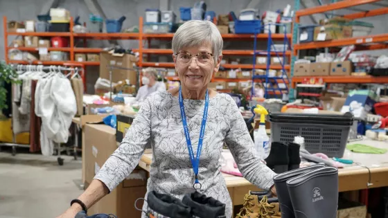 Woman with short silver hair and glasses stands behind a tray of boots and shoes. In the background is a warehouse with clothing and other items.